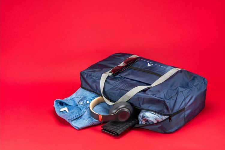 Creative lifestyle shot of a half opened travel bag in blue color revealing the items of the bag including shirt, headphones and purse on a red background by Isa Aydin NJ NY LA
