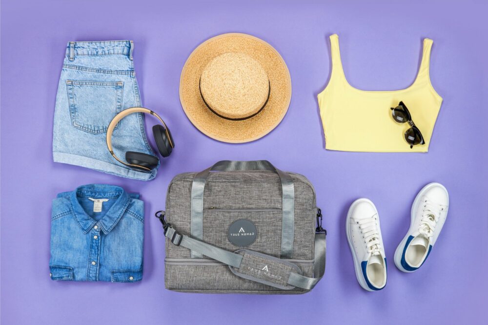 Creative lifestyle photography of a travel bag with necessary travelling items like hat, sunglasses, trouser, shirt, blouse and shoes on a purple background by Isa Aydin NJ NY LA