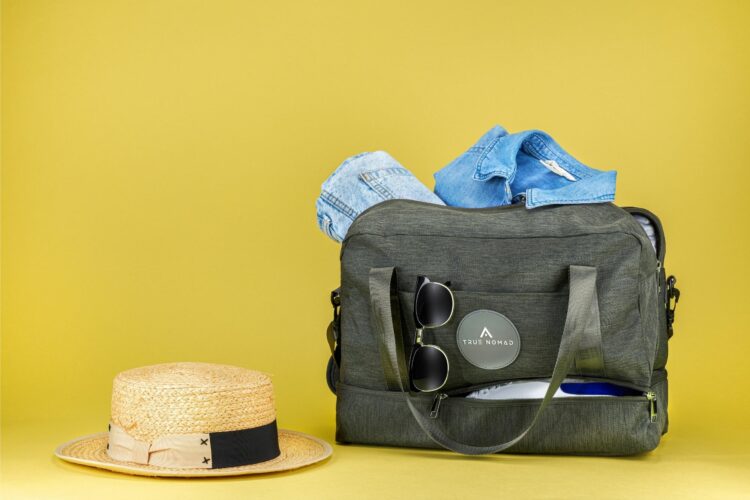 Creative shot of a voyager colored travel bag revealing the items placed inside the bag including sunglasses, shirt, jeans and shoes while a hat is placed on the side.