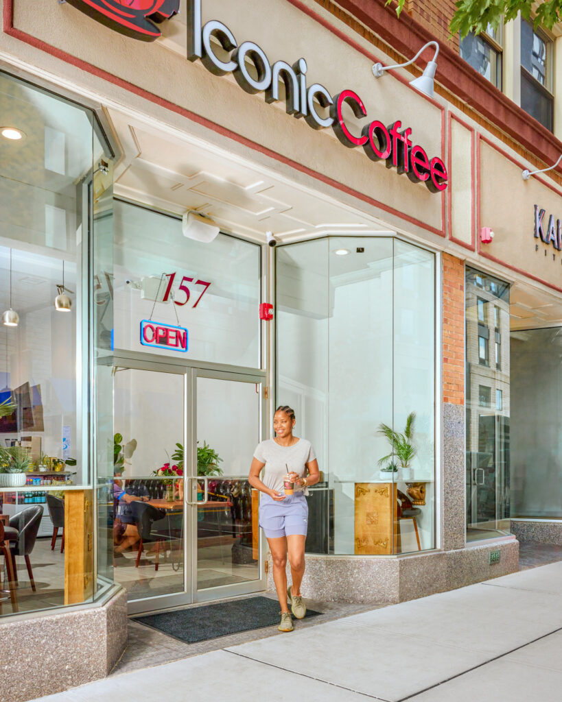 A smiling woman exits Iconic Coffee shop at 157 Main Street, carrying a cold beverage. Above her, the shop's red cursive 'Iconic Coffee' sign stands out, with a bright 'Open' neon sign visible through the glass facade. Inside, patrons enjoy the shop's cozy ambiance. This image is a part of Isa Aydin's editorial photography portfolio, showcasing a vibrant street scene and commercial business in operation