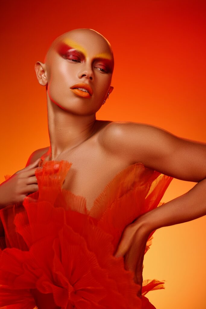 A fashion editorial portrait of a model exuding confidence with a shaved head, dramatic red and yellow makeup, and vibrant red lips. The model is dressed in an avant-garde red ruffled outfit, striking a poised pose against a saturated orange backdrop, epitomizing the bold and transformative essence of high fashion."