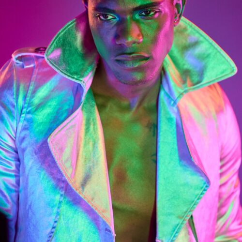 Fashion editorial portrait of a model in a dynamic pose, illuminated by dramatic green and pink lighting. The model's expression is intense and focused, with a sharp gaze into the camera. He is dressed in a futuristic, metallic silver coat with an upturned collar that reflects the vivid lighting, creating a holographic effect on the fabric. The background's deep purple and pink shades enhance the avant-garde and stylish atmosphere of the photo, typical of high-end fashion photography.