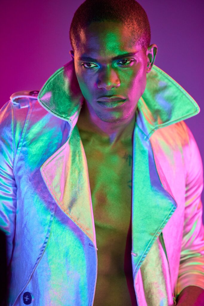 Fashion editorial portrait of a model in a dynamic pose, illuminated by dramatic green and pink lighting. The model's expression is intense and focused, with a sharp gaze into the camera. He is dressed in a futuristic, metallic silver coat with an upturned collar that reflects the vivid lighting, creating a holographic effect on the fabric. The background's deep purple and pink shades enhance the avant-garde and stylish atmosphere of the photo, typical of high-end fashion photography.