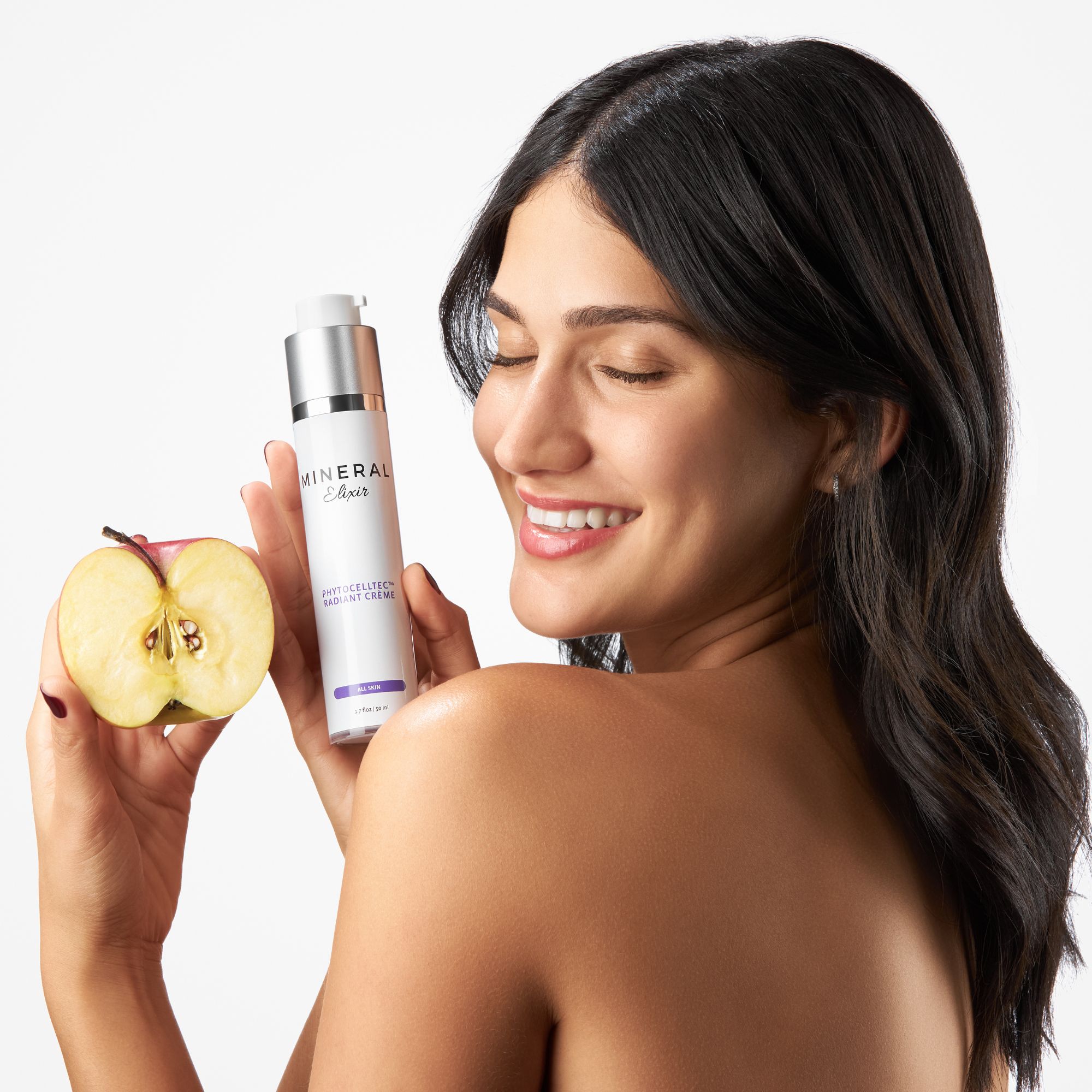 Smiling woman holding a fresh apple in one hand and a bottle of Mineral Enriched Radiant Cream in the other, showcasing natural skincare products on a white background
