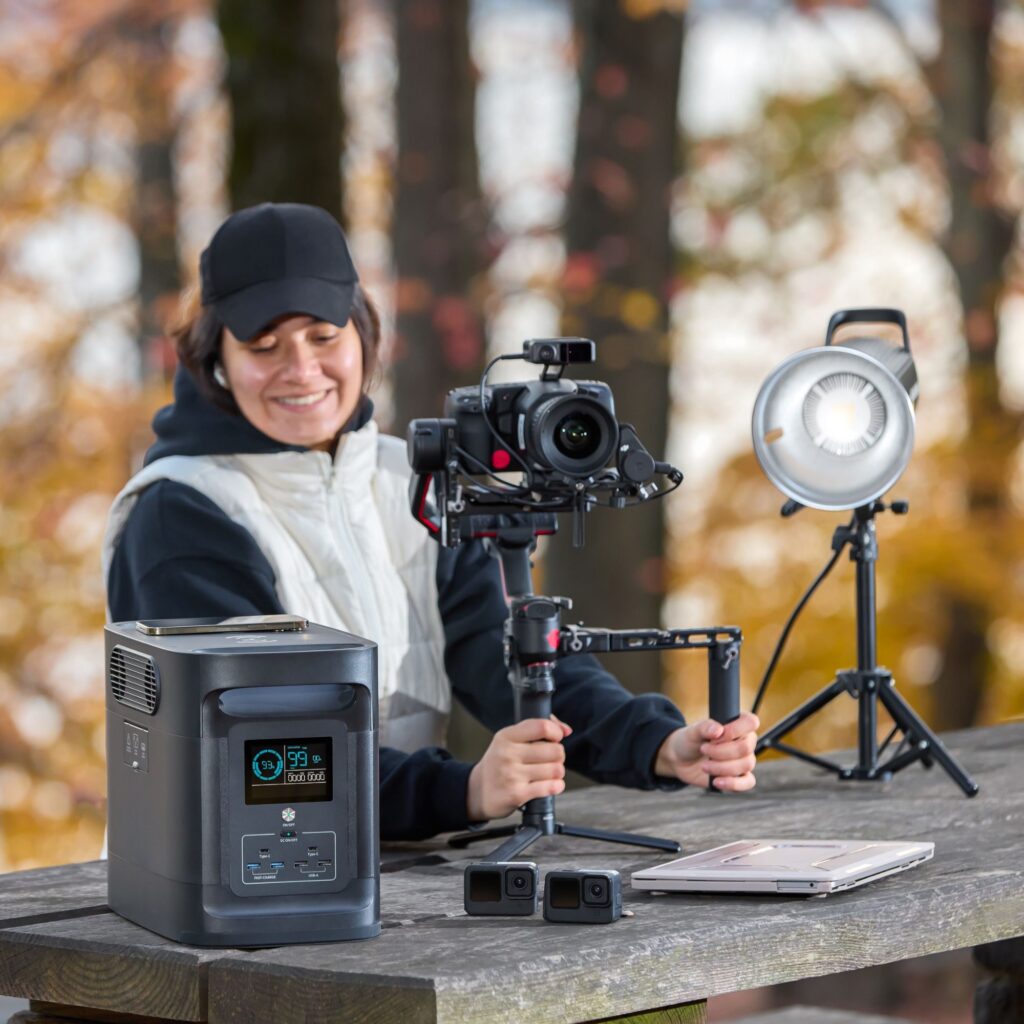 A smiling woman operating a camera on a gimbal with a LED light panel and a portable solar power bank generator displayed on a wooden table, illustrating a professional product photography setup in an outdoor environment.