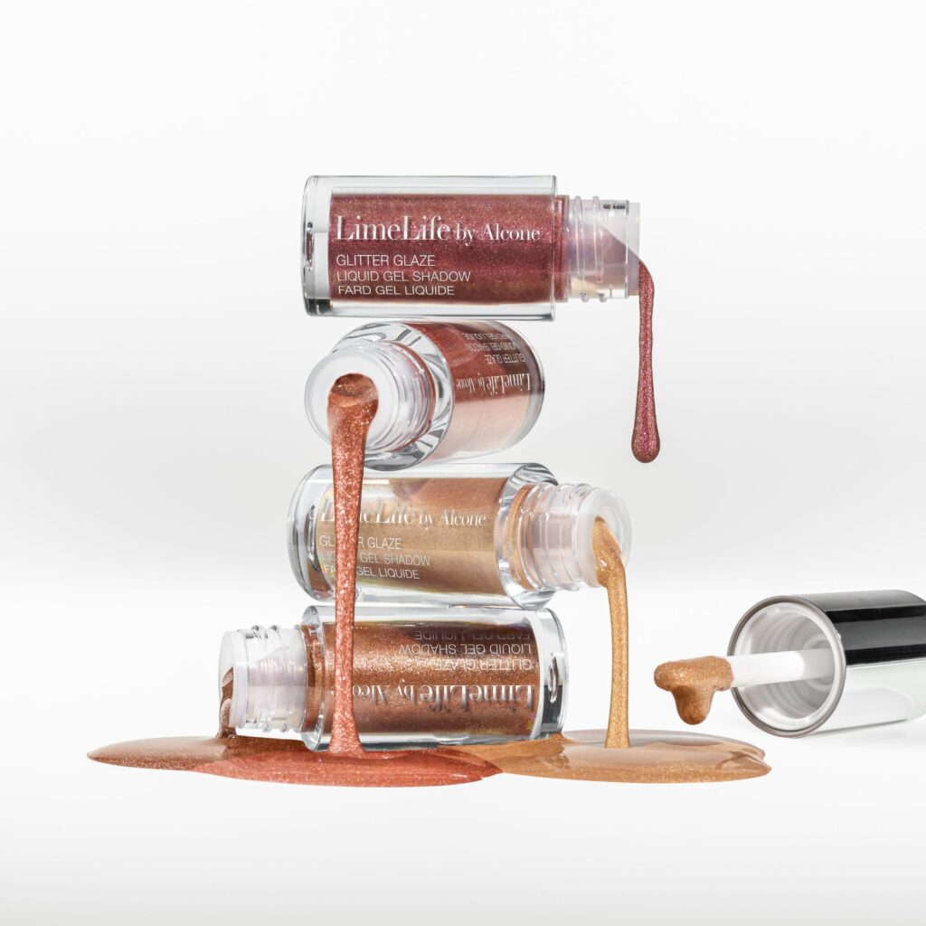 liquid eye shadow photography of A balanced stack of three 'LimeLife by Alcone' liquid gel shadow pots in shades of rose, bronze, and gold. The pots are open with the product spilling over the edges, creating artistic drips and pools on the white surface. One of the applicators is lying next to the pots, covered in the golden product.